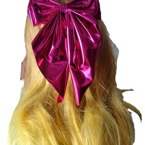 May include: A large, shiny magenta bow hair accessory. The bow is made of a metallic material and is attached to blonde hair. The bow is centered on the head and is a prominent feature of the image.