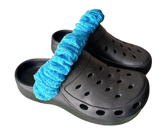 May include: A pair of black clogs with a blue lace shoe accessory. The clogs have multiple oval-shaped holes and a thick sole. The blue accessory is a ruffled lace band.