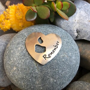 May include: A heart-shaped metal token with a brushed finish featuring a cutout of a candle flame and the word Remember stamped in black ink. The token is resting on a smooth gray stone, positioned among other stones and a small succulent plant.