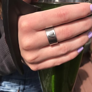 May include: A silver ring with a heart design. The ring is worn on a finger with light purple nail polish.