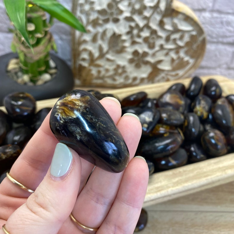 May include: A close-up of a dark amber gemstone with brown and black inclusions. The gemstone is smooth and polished, and it is being held in a hand.