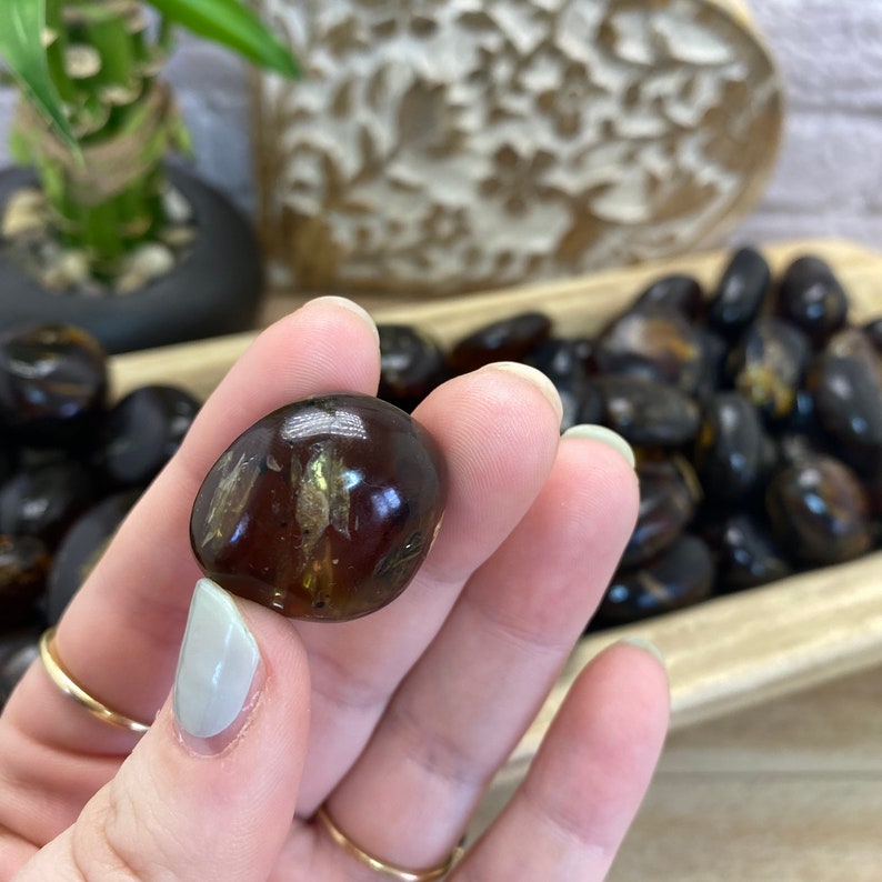 May include: A close-up of a dark brown tumbled stone with a lighter brown, translucent center. The stone is being held in a hand.