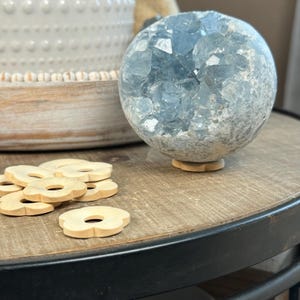 May include: A large, blue celestite crystal sphere on a wooden flower-shaped stand. The sphere is surrounded by a light-coloured, rough exterior. Several small, wooden flower-shaped cutouts are in the foreground. The sphere sits on a dark wooden table.