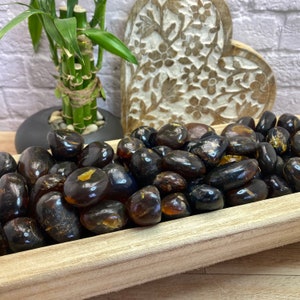 May include: A wooden bowl filled with dark amber tumbled stones. The stones are smooth and polished, with a variety of shapes and sizes.