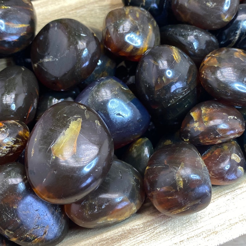 May include: A close-up of a collection of tumbled amber stones. The stones are a deep brown color with hints of gold and yellow. They are smooth and polished, with a glossy finish.
