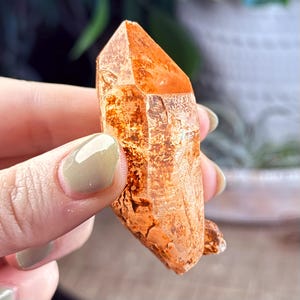 Puede incluir: Un gran cristal de cuarzo transparente naranja con una textura natural rugosa. El cristal es puntiagudo en un extremo y tiene una base plana.
