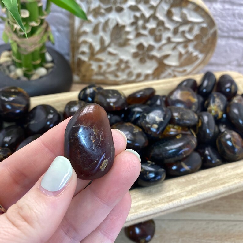 May include: A close-up of a dark amber tumbled stone. The stone is smooth and polished, with a slightly reddish hue. The stone is being held in a hand, and there are other tumbled stones in the background.