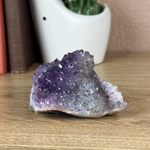 Puede incluir: Un gran cúmulo de cristal de amatista de color morado y gris. El cristal tiene una textura rugosa y natural.