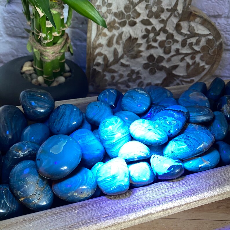 May include: A collection of smooth, blue and white tumbled stones in a wooden tray. The stones have a swirling pattern and are illuminated by a blue light.