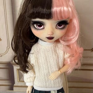 Op de afbeelding: Modepop met half bruin en half roze haar, gekleed in een witte trui en een donkergroene rok met een wit bloemmotief. De pop heeft grote ogen en donkere lippenstift.