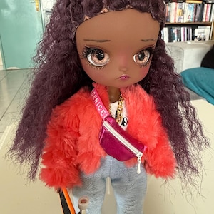 Peut inclure: Poupée de mode à la peau foncée, vêtue d'une veste rouge vif et floue, d'un jean bleu clair et d'un sac banane rose. La poupée a des cheveux violets et tient un skateboard orange. Un collier en chaîne dorée est visible.