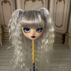 Könnte beinhalten: Ein Puppenkopf mit heller Haut, großen blauen Augen und langen, silbergrauen Haaren, die zu Zöpfen gestylt sind. Die Puppe hat dunkelrote Lippen und lange Wimpern. Der Kopf ist auf einem Holzstab mit einem dekorativen blau-weißen Blumenmuster befestigt.