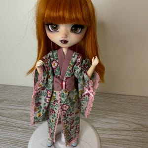 May include: A doll with auburn hair and a floral kimono. The kimono is green with pink and white flowers, trimmed with pink lace. The doll has large eyes and is standing on a white base.
