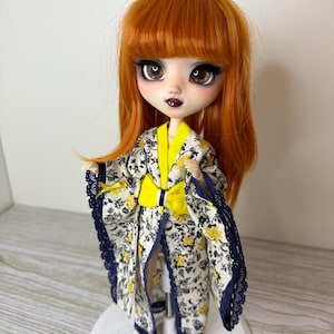 Può includere: Una bambola con capelli rossi e un kimono floreale. Il kimono è bianco con motivi floreali gialli, un fiocco giallo e finiture in pizzo blu navy. La bambola ha grandi occhi e si trova su una base bianca.