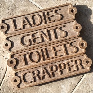 May include: Four wooden signs with rounded edges and holes in the corners. The signs are stacked on top of each other and read "LADIES", "GENTS", "TOILETS", and "CRAPPER".