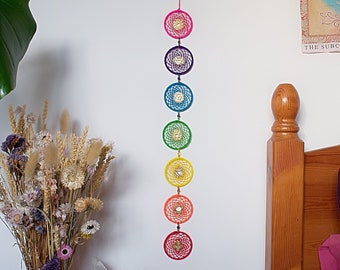 May include: A rainbow colored dreamcatcher wall hanging with seven circular hoops, each with a different color of yarn. The dreamcatcher is hanging on a white wall with a wooden headboard and a plant in the foreground.