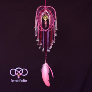 Op de afbeelding: Een roze en paarse droomvanger met een kristallen hanger en een roze veer die eraan hangt. De droomvanger is versierd met kralen en kristallen.