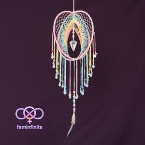 May include: A heart-shaped dreamcatcher with a rainbow design. The dreamcatcher features a central crystal pendant and is adorned with colorful beads and hanging crystal droplets. A feather dangles from the bottom. The logo for feminfinite is in the lower left corner.