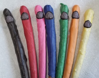 May include: Eight pretzel rods dipped in different colored chocolate. The chocolate is black, red, pink, blue, green, orange, and yellow. Each chocolate-covered pretzel has a small gray chocolate piece on top.