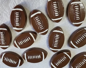 Op de afbeelding: Een bovenaanzicht van verschillende chocolade American footballs. Elke bal is bruin met witte strepen en veters. De ballen liggen op een wit, getextureerd oppervlak.