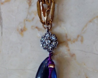 Puede incluir: Un collar de cadena dorada con un colgante de piedra azul en forma de lágrima. El colgante está acentuado con un pequeño grupo de diamantes de imitación en forma de flor.