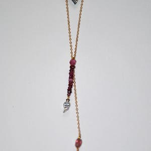 Peut inclure: Un collier en chaîne dorée avec un petit pendentif triangulaire argenté serti de pierres claires. Le collier a deux brins, l'un avec un groupe de pierres roses et un petit pendentif en forme de feuille argenté serti de pierres claires, et l'autre avec une seule pierre rose et un pompon rose.