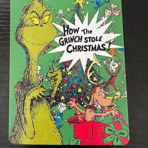 以下が含まれることがあります： 「How the Grinch Stole Christmas」の緑色のカード。 グリンチはいたずらっぽい笑顔で描かれ、カラフルなキャラクターと装飾されたクリスマスツリーに囲まれています。 タイトルは白い星の中に表示されています。