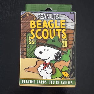 May include: A deck of Peanuts Beagle Scouts playing cards. The box features Snoopy in a scout uniform, with the words "BEAGLE SCOUTS" in large yellow letters. The background shows a cabin and trees.