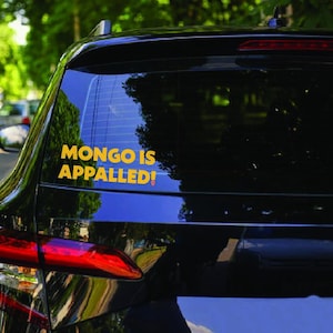 May include: A black car rear window featuring a yellow vinyl decal with the text "MONGO IS APPALLED!" in a bold, sans-serif font. The sticker is applied to the lower left section of the glass, contrasting against the dark, reflective surface.