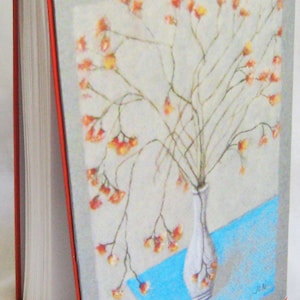 May include: A handmade journal with a red cover and a gray spine. The cover features a painting of a vase with dried flowers in shades of orange and yellow. The vase is white and sits on a blue surface. The artist's initials "J.M." are in the bottom right corner of the painting.