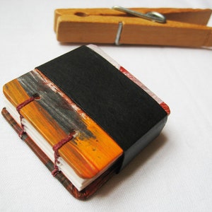 Pu&ograve; includere: Un piccolo libro fatto a mano con una copertina in pelle nera e pagine dipinte di arancione, giallo e rosso. Il libro &egrave; chiuso e ha un nastro rosso legato intorno.