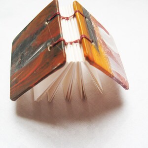 Pu&ograve; includere: Un piccolo libro fatto a mano con copertine dipinte. Le copertine sono un mix di vernice rossa, arancione, marrone e nera. Il libro &egrave; chiuso e le pagine sono visibili.