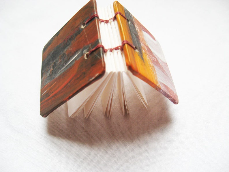 Pu&ograve; includere: Un piccolo libro fatto a mano con copertine dipinte. Le copertine sono un mix di vernice rossa, arancione, marrone e nera. Il libro &egrave; chiuso e le pagine sono visibili.