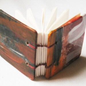 Pu&ograve; includere: Un piccolo libro fatto a mano con una copertina dipinta astratta e colorata. La copertina &egrave; un mix di tonalit&agrave; rosse, arancioni, gialle e blu. Il libro &egrave; chiuso e le pagine sono visibili.
