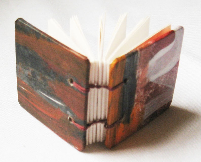 Pu&ograve; includere: Un piccolo libro fatto a mano con una copertina dipinta astratta e colorata. La copertina &egrave; un mix di tonalit&agrave; rosse, arancioni, gialle e blu. Il libro &egrave; chiuso e le pagine sono visibili.