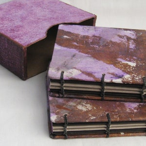 May include: Two small, handmade journals with purple and brown covers. The journals are bound with black thread and have a layered look. The journals are sitting on a purple box.
