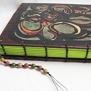 May include: A handmade journal with a dark brown cover featuring a black and white abstract design. The journal has a green spine with black stitching and a brown tassel.
