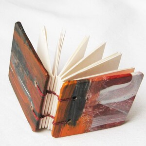 Pu&ograve; includere: Un piccolo libro fatto a mano con copertine dipinte colorate. Il libro &egrave; chiuso e le pagine sono a ventaglio. Le copertine sono dipinte in tonalit&agrave; di arancione, rosso, marrone e nero.