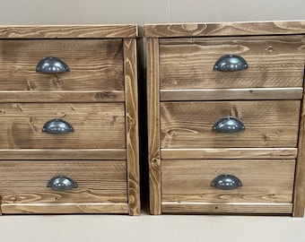 Puede incluir: Dos mesitas de noche de madera, cada una con tres cajones. Las mesitas están hechas de madera marrón claro con vetas visibles y tienen tiradores oscuros de metal en forma de taza. El diseño es rústico y sencillo, adecuado para un dormitorio.