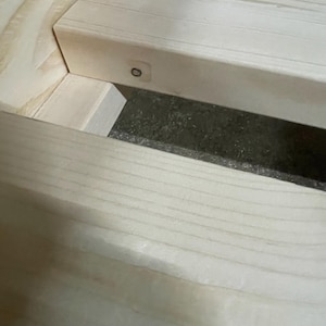 Op de afbeelding: Een lichtgekleurd houten bedframe met horizontale latten verbonden aan een zijbalk. De natuurlijke houtnerf is zichtbaar over het lichtbeige oppervlak, met een kleine metalen schroef zichtbaar aan de zijkant van de houten lat.