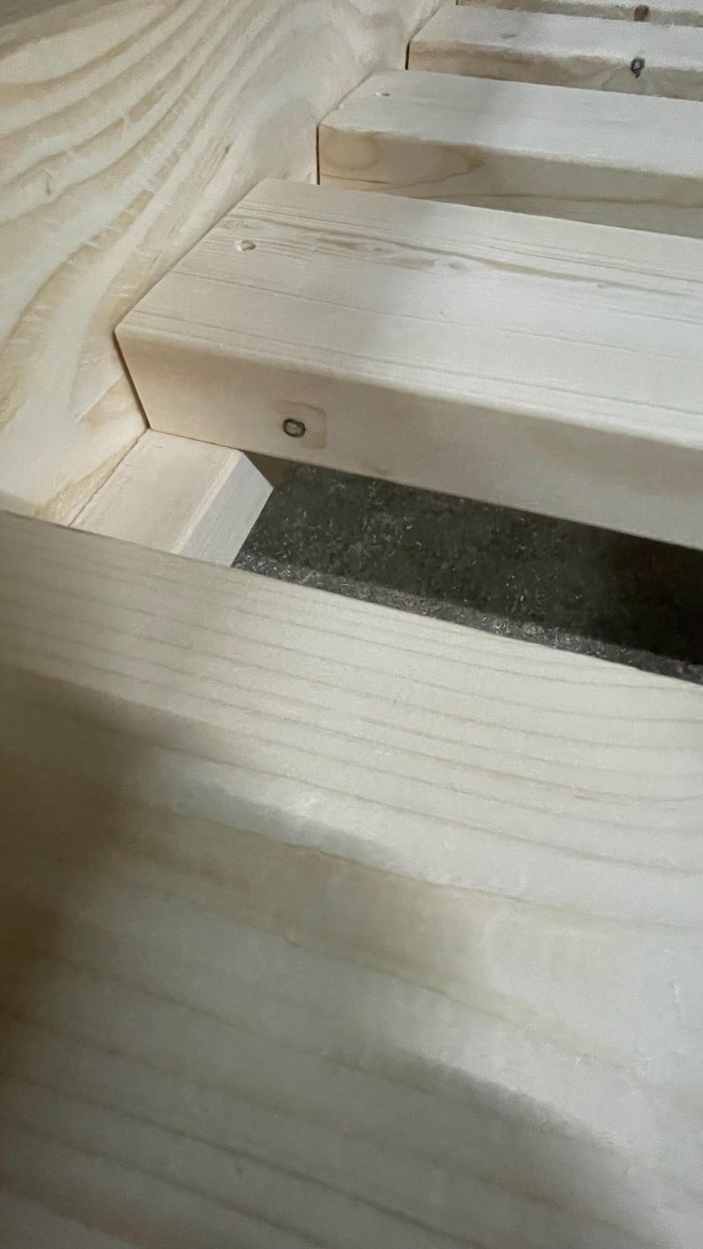 Op de afbeelding: Een lichtgekleurd houten bedframe met horizontale latten verbonden aan een zijbalk. De natuurlijke houtnerf is zichtbaar over het lichtbeige oppervlak, met een kleine metalen schroef zichtbaar aan de zijkant van de houten lat.