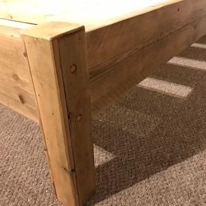 Op de afbeelding: Een lichtbruin houten bedframe met een stevige hoekpaal met zichtbare ronde deuvelverbindingen. Het frame is opgebouwd uit planken met een natuurlijke houtnerftextuur en toont een eenvoudig, functioneel ontwerp als matrasbasis.