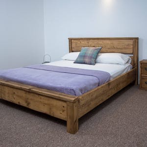May include: A rustic wooden bed frame with a light brown finish. The bed is made with reclaimed wood and has a simple, modern design. The bed is dressed with a purple blanket and white pillows.
