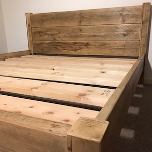 Op de afbeelding: Een houten bedframe met een hoog hoofdeinde opgebouwd uit horizontale planken met een verweerde afwerking. De basis bestaat uit lichtgekleurde, onbewerkte houten latten die horizontaal zijn geplaatst om een matras te ondersteunen.