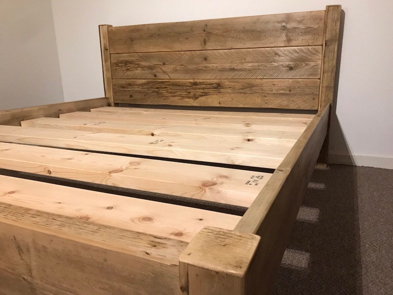 Op de afbeelding: Een houten bedframe met een hoog hoofdeinde opgebouwd uit horizontale planken met een verweerde afwerking. De basis bestaat uit lichtgekleurde, onbewerkte houten latten die horizontaal zijn geplaatst om een matras te ondersteunen.