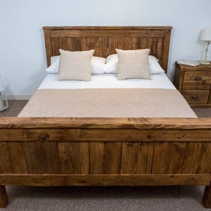 Puede incluir: Un marco de cama de madera r&uacute;stica con un acabado de madera natural. La cama tiene una cabecera y un piecero con un dise&ntilde;o de listones. La cama est&aacute; hecha de madera recuperada y tiene un aspecto desgastado.