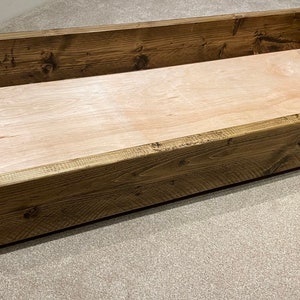 May include: A rectangular wooden storage box with a light-coloured interior and dark brown exterior. The box is on small black wheels and appears to be made of solid wood. The box is approximately 100 cm long, 50 cm wide, and 25 cm high.