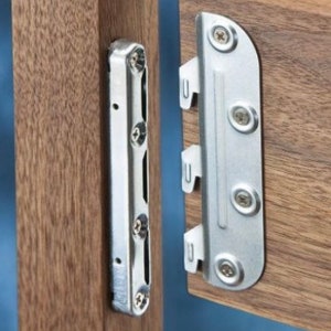 Puede incluir: Primer plano de un soporte met&aacute;lico con cuatro tornillos, fijado a una superficie de madera. El soporte est&aacute; dise&ntilde;ado para conectar dos piezas de madera.
