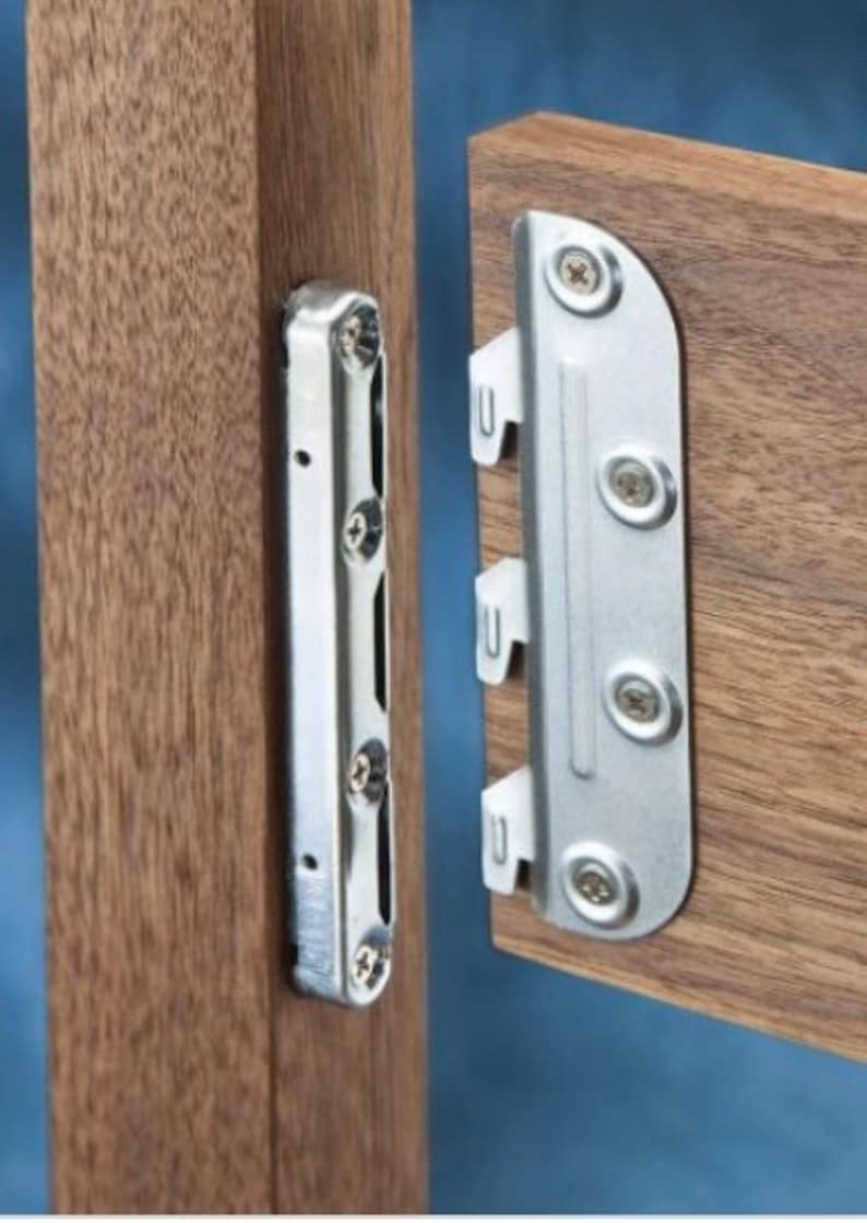 Puede incluir: Primer plano de un soporte met&aacute;lico con cuatro tornillos, fijado a una superficie de madera. El soporte est&aacute; dise&ntilde;ado para conectar dos piezas de madera.