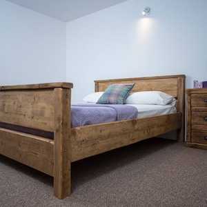 May include: A rustic wooden bed frame with a light brown finish. The bed has a headboard, footboard, and side rails. The bed is made of reclaimed wood and has a natural, distressed look. The bed is dressed with a purple blanket and a plaid pillow.